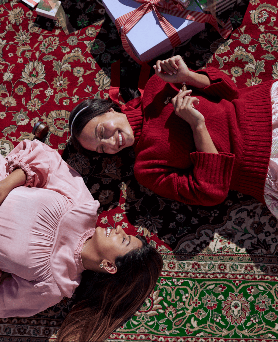 Two young women lay on a carpet on their backs smiling at one another.