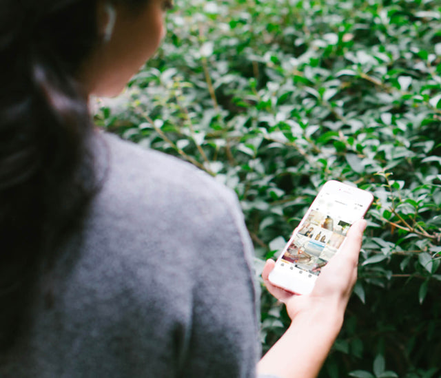 A person with long dark hair holds an iPhone against a leafy green backdrop.