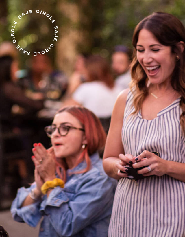 A group of joyful, clapping women surrounded by greenery sit at a table with a brunette woman standing and laughing beside.