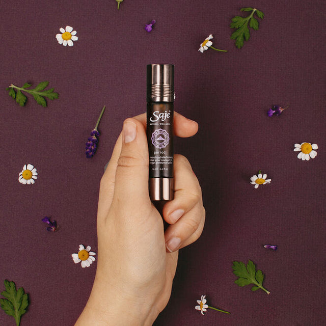 A hand holding the Period pre-menstrual relief remedy roll-on with cap on with roman chamomile, geranium and lavender behind on a plum background